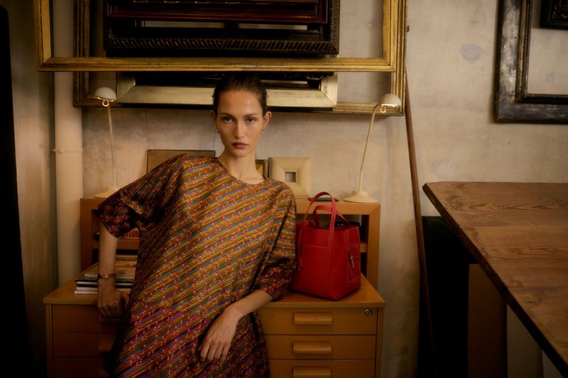Model wearing patterned dress leaning on a wooden cabinet next to a bright red handbag in an interior setting.