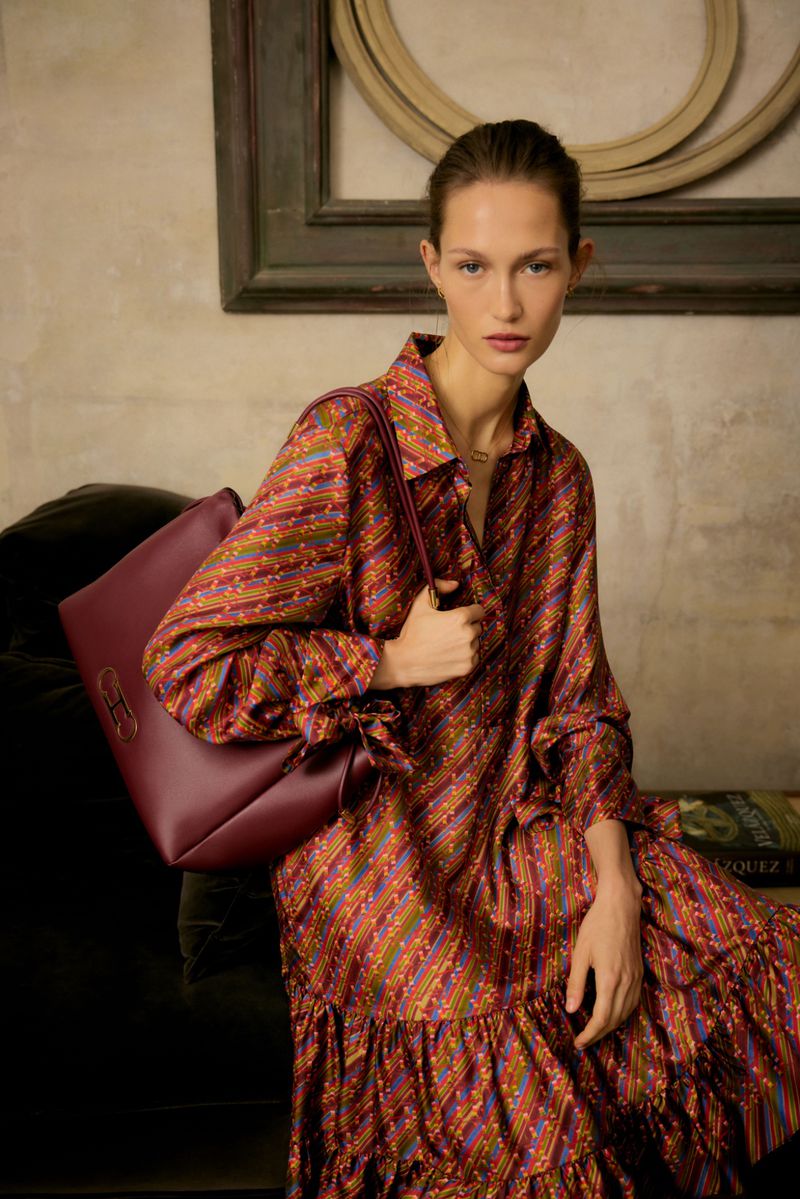 Model in patterned blouse holding burgundy leather handbag against neutral interior