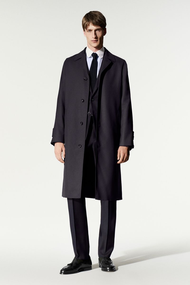 Model in dark overcoat and suit with a black tie standing against a white background