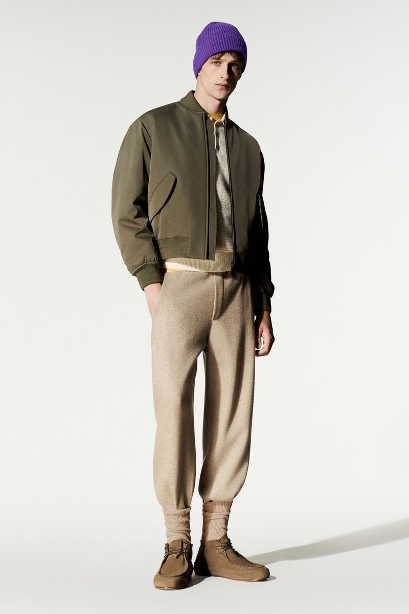 Model wearing an olive green bomber jacket, knit beige trousers, and a purple beanie against a white studio background.