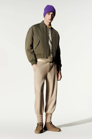 Model wearing an olive green bomber jacket, knit beige trousers, and a purple beanie against a white studio background.