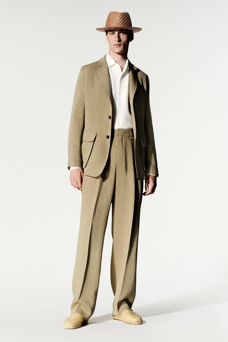 Model wearing a tan suit, white shirt, straw hat, and light shoes against a seamless white studio backdrop