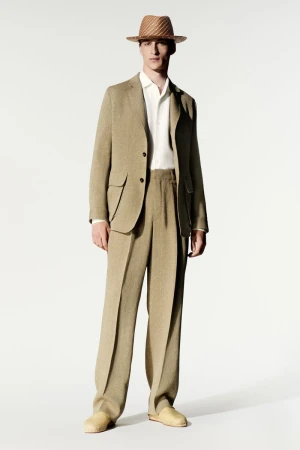 Model wearing a tan suit, white shirt, straw hat, and light shoes against a seamless white studio backdrop