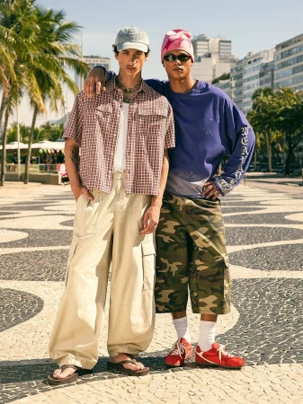 Two male models posing outdoors on a tiled promenade with palm trees and buildings in the background for Bershka