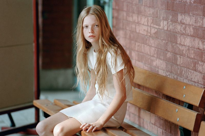 Young girl with long blonde hair wearing a white t-shirt and skirt seated on a wooden bench against a red brick wall