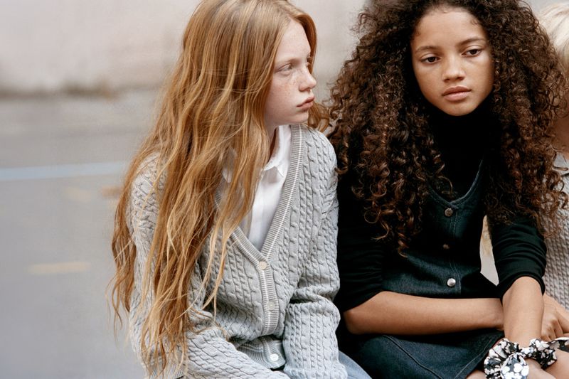 Two young female models sitting outdoors, wearing a grey cable knit cardigan and a dark dress for Dior Kids.