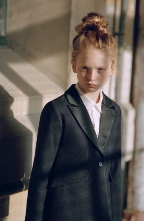 Young child in a dark tailored blazer and white shirt with hair in a high bun standing in natural sunlight