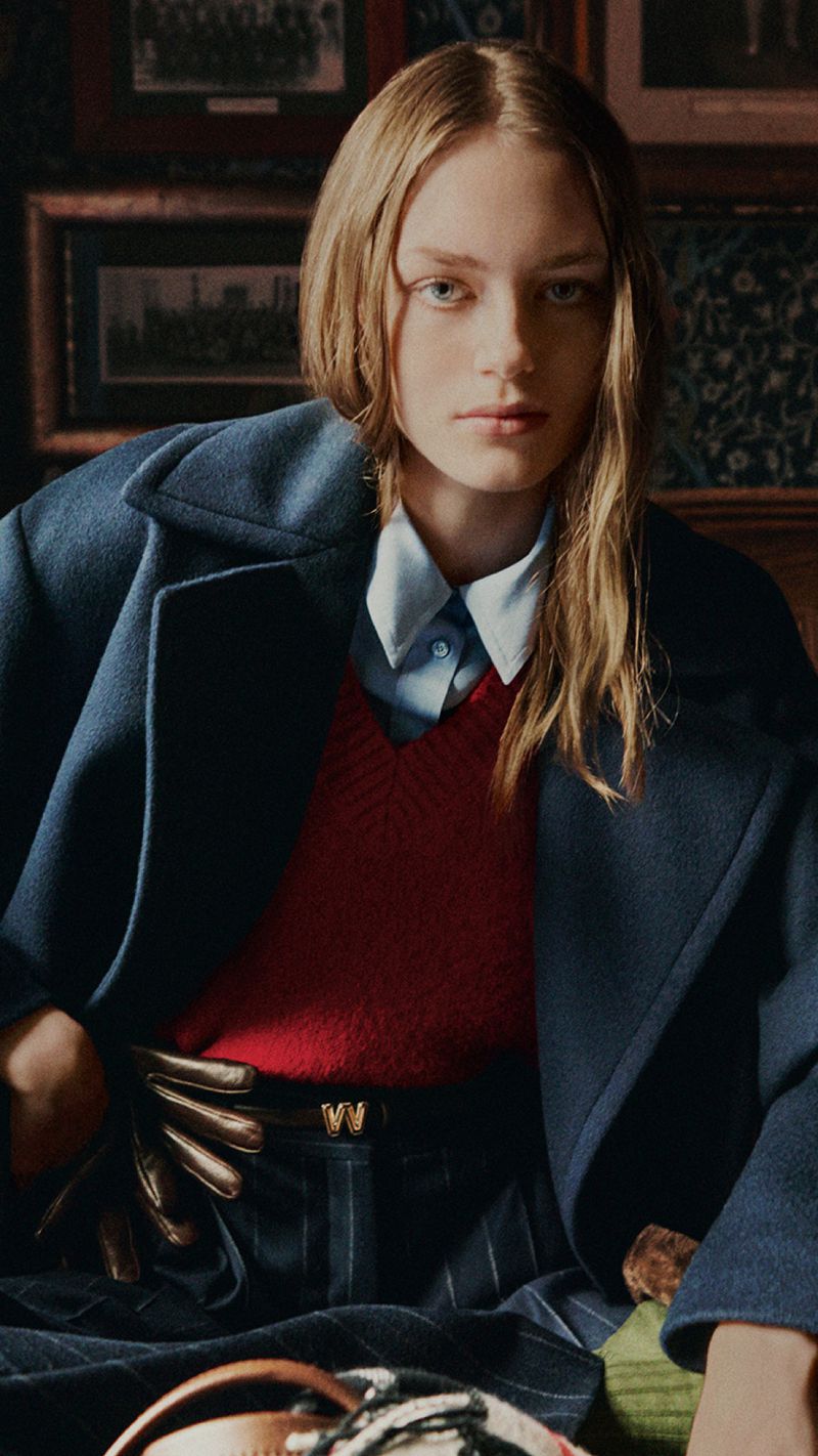 A female model wearing a navy coat, red vest, and collared shirt indoors against a dark framed wall.