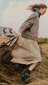 Model walking through grassy dunes holding a brown leather tote bag wearing a patterned jacket and beige skirt