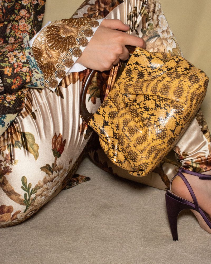 Close-up of a person holding a yellow snakeskin handbag while wearing floral satin trousers and heels for Heroine Magazine.