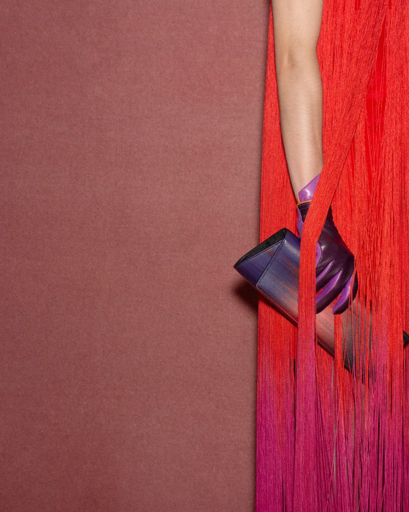 Close-up of a hand in a purple glove holding a sleek handbag against a red fringed dress and textured background