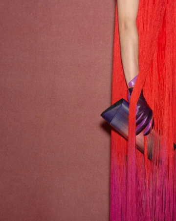 Close-up of a hand in a purple glove holding a sleek handbag against a red fringed dress and textured background