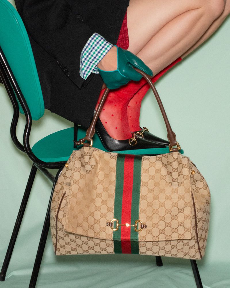 Model seated on a turquoise chair holding a canvas handbag with red and green stripes, wearing green leather gloves.