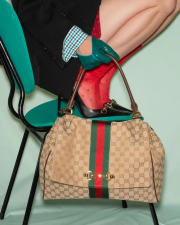 Model seated on a turquoise chair holding a canvas handbag with red and green stripes, wearing green leather gloves.