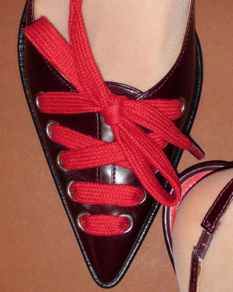 Close-up of a glossy dark red pointed shoe laced with thick red laces against a solid brown background.
