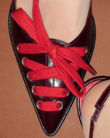 Close-up of a glossy dark red pointed shoe laced with thick red laces against a solid brown background.