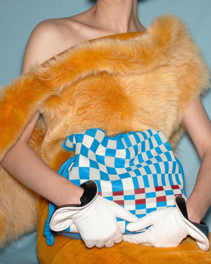 Model wearing a fuzzy orange stole and white leather gloves holding a blue and white checkered handbag.