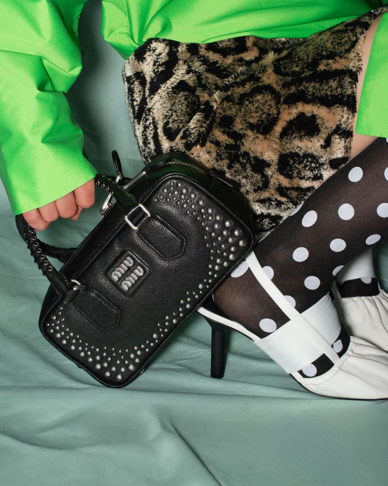 Close-up fashion editorial shot of a black studded leather handbag held by a person in a green jacket and leopard print skirt.