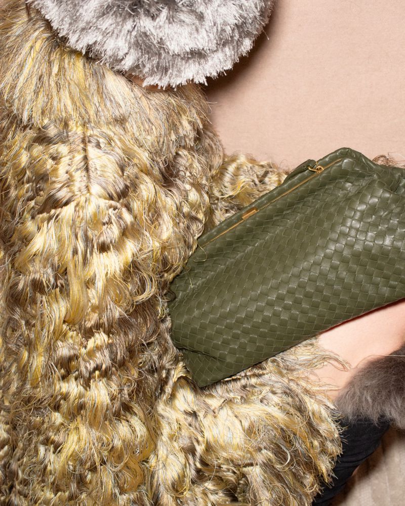 Close-up of a textured shaggy coat and an olive green woven leather clutch bag against a plain background.