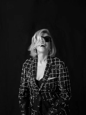 Portrait of Christine Baranski laughing in sunglasses and a grid-patterned blazer against a dark background.