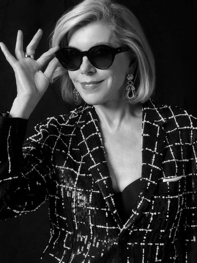 Portrait of Christine Baranski wearing dark sunglasses and a patterned blazer against a dark background