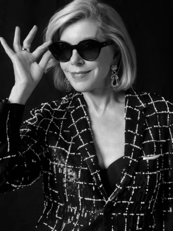 Portrait of Christine Baranski wearing dark sunglasses and a patterned blazer against a dark background