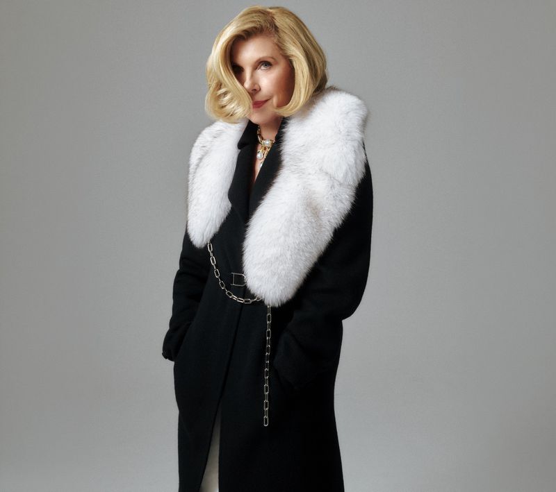 Christine Baranski wearing a black coat with a white fur collar against a grey background