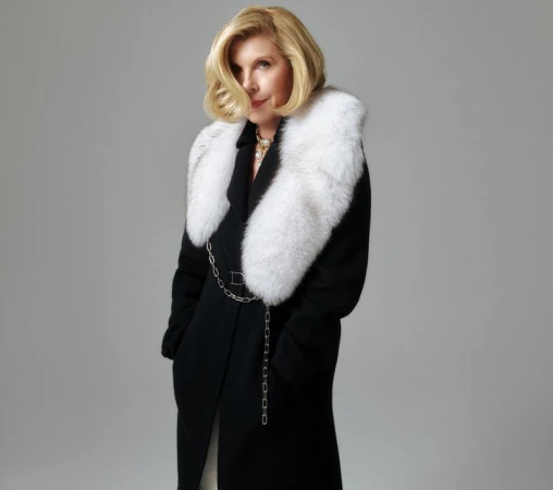 Christine Baranski wearing a black coat with a white fur collar against a grey background