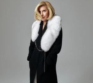 Christine Baranski wearing a black coat with a white fur collar against a grey background