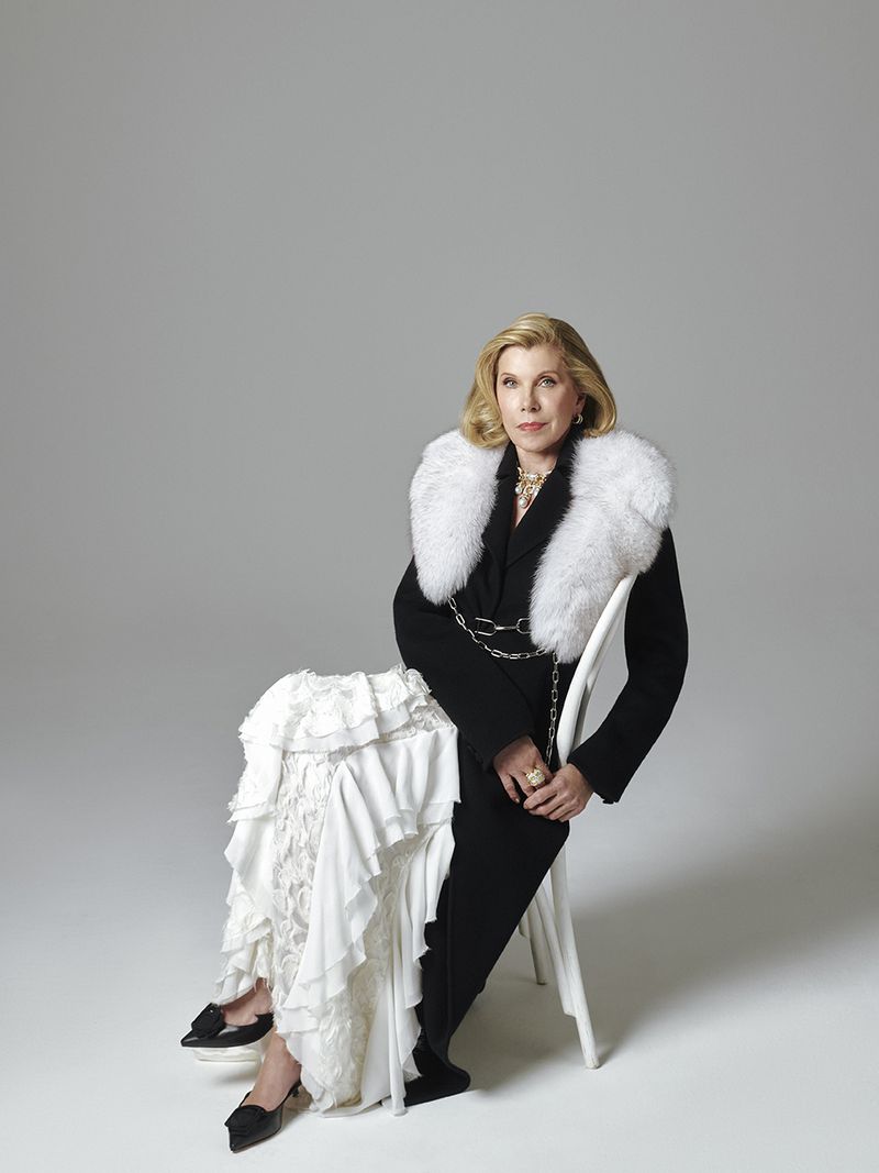 Christine Baranski seated on a white chair wearing a black coat with a white fur collar and ruffled white skirt against a light grey background