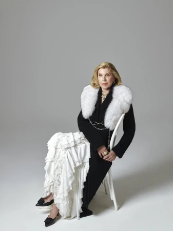 Christine Baranski seated on a white chair wearing a black coat with a white fur collar and ruffled white skirt against a light grey background