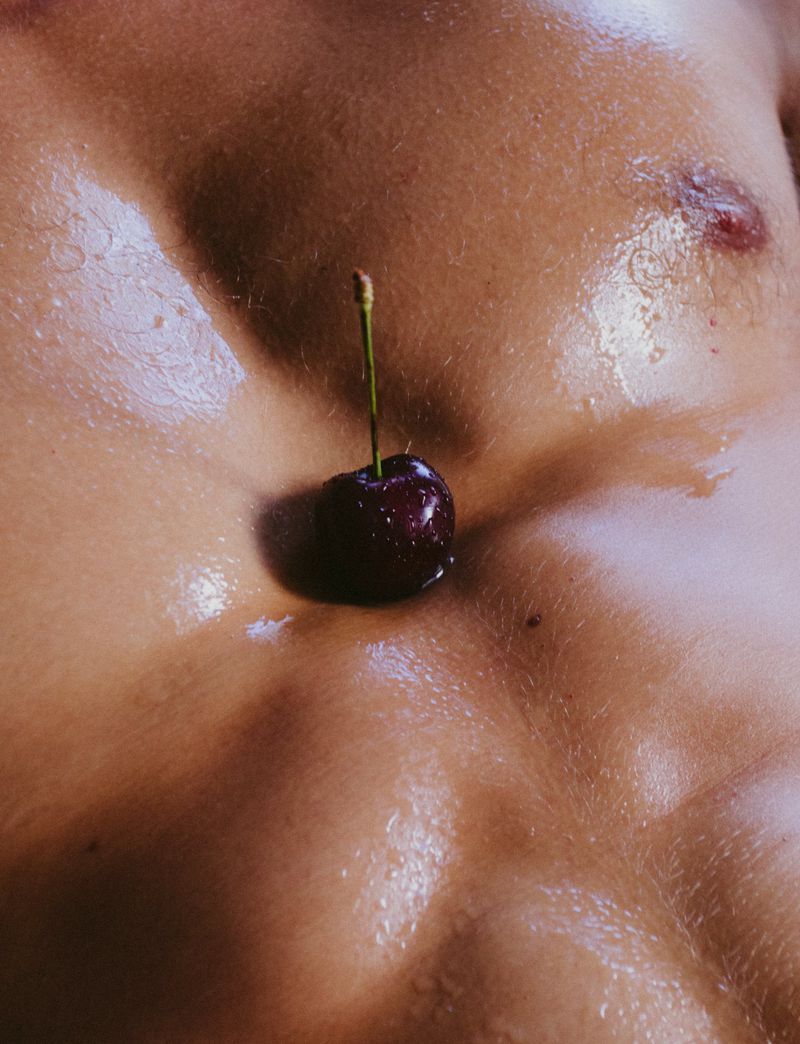 Close-up of a single cherry on an adult male's chest, highlighting the contrast between the fruit's deep red and the skin's texture.