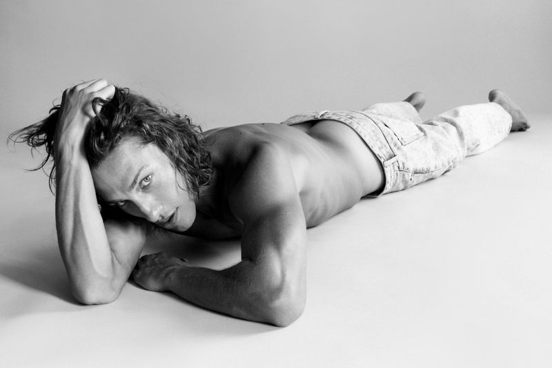 Black and white studio shot of a shirtless man lying on his stomach, looking into the camera, with his hand on his head.