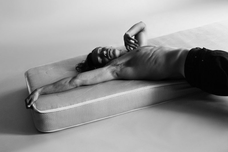 Monochrome studio shot of an adult male lying on a mattress, arm outstretched, eyes closed, against a pale grey backdrop.
