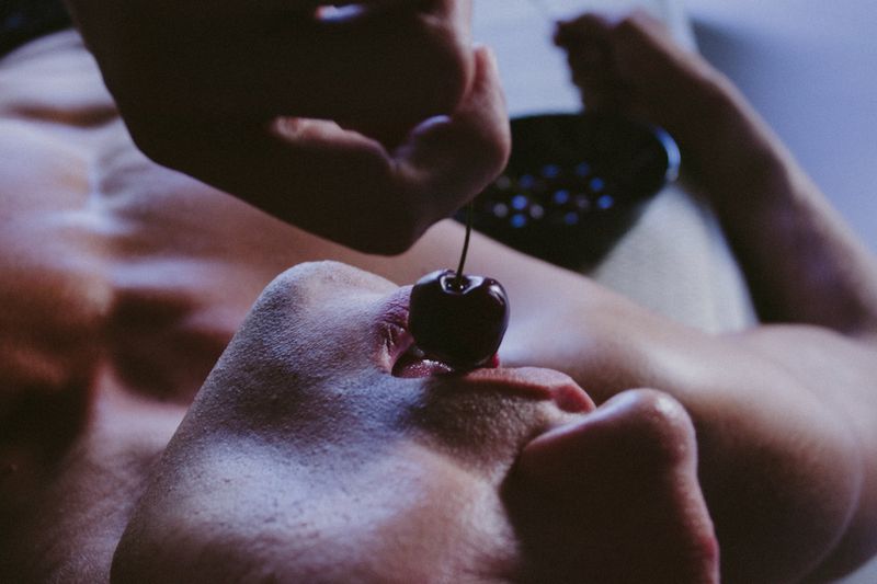Man lying with a cherry resting on his lip, held in place by a hand above, with a black colander in the blurred background.