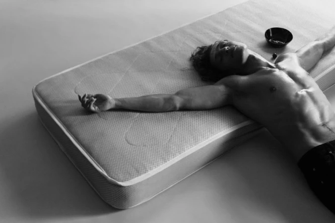 Muscular man with curly hair lies shirtless on a mattress in a photo studio in black and white with a dark bowl nearby.