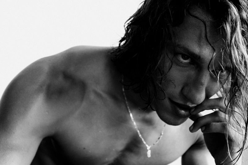 Close-up black and white portrait of an adult male with long dark hair, shirtless with a silver chain, against a white background.