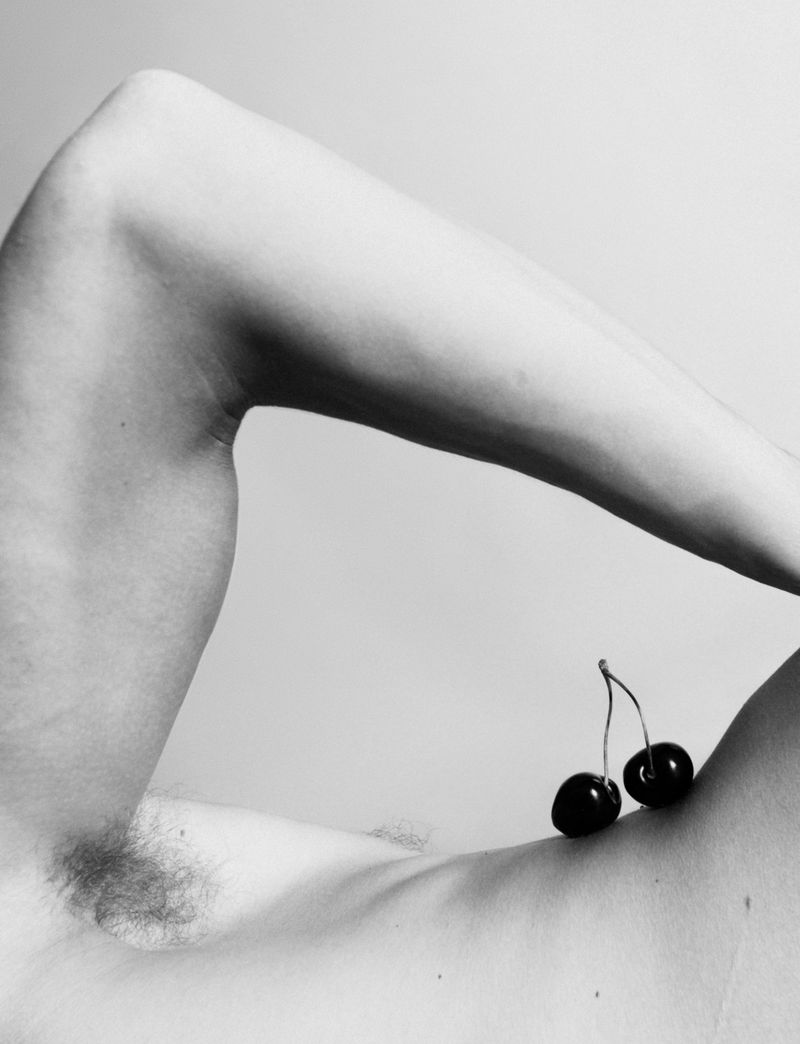 Black and white nude torso of a man with two cherries resting on his chest, arm angled above. Simple background, no distractions.