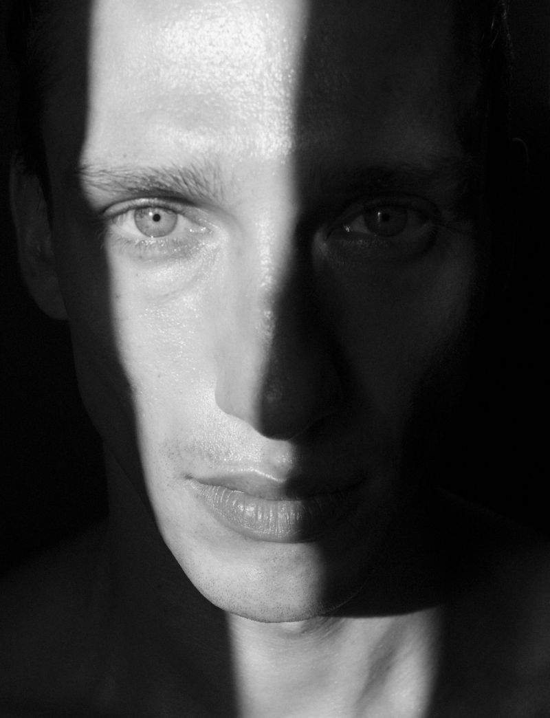 Black and white portrait of an adult male with a stripe of light dramatically illuminating half of his face in a high-contrast image.