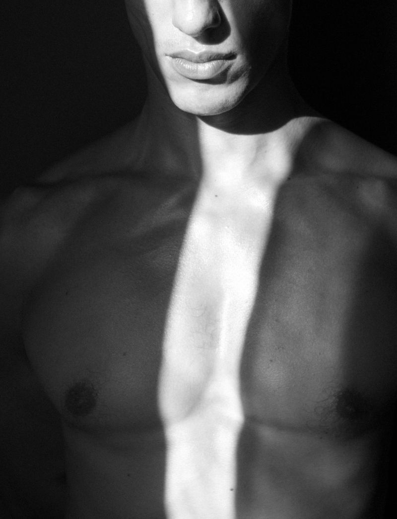 Monochrome close-up photo of a shirtless adult male with a light stripe highlighting his chest and upper torso against a dark background.