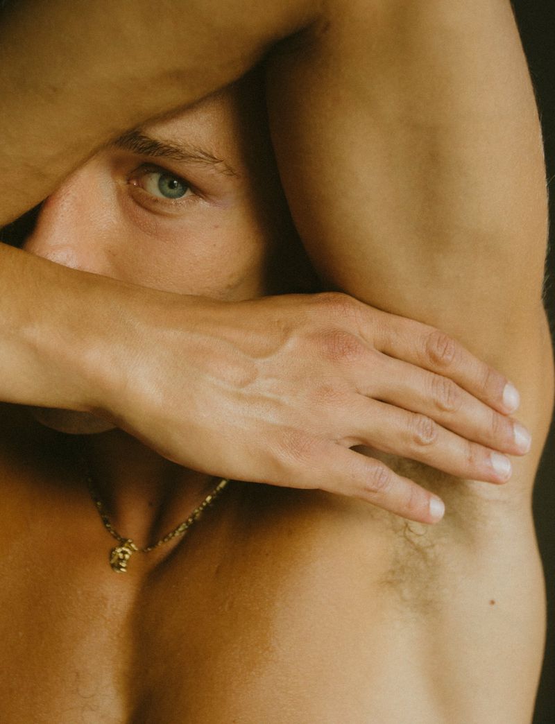 Close-up shot of a shirtless adult male with blue eyes, fair skin, arm covering his face, and wearing a gold chain necklace.