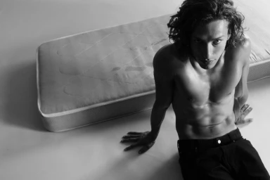 Monochrome portrait of a shirtless man with long curly hair seated on the floor beside a mattress.