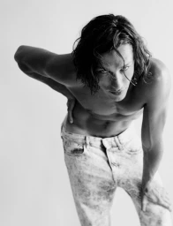 Black and white portrait of a shirtless man with long dark hair wearing light patterned jeans against a plain backdrop.