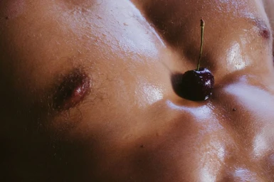 Close-up of a glistening muscular male torso with a dark cherry resting in the navel.