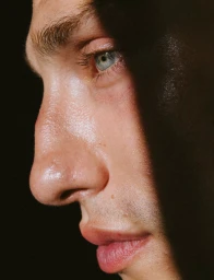 Close-up profile portrait of a man's face with bright blue eyes and textured skin in dramatic low-key lighting against a dark background.