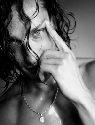 Black and white close up portrait of a young man with wet dark hair touching his eyebrow and wearing a silver pendant necklace.