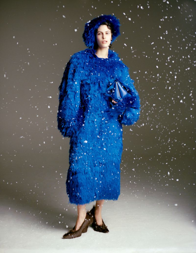 Model in vibrant blue fluffy coat and matching hat standing against a pale grey studio background amidst falling snow