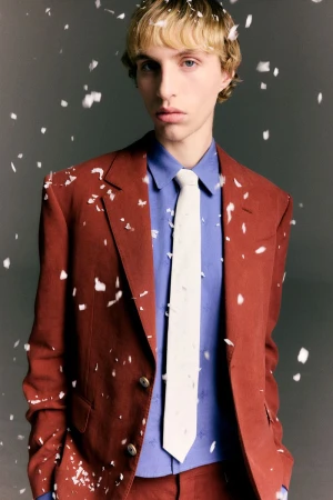 Model in rust-brown suit jacket with a blue shirt and white tie amid falling snow or confetti flakes