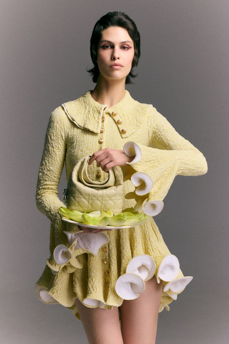Model wearing a pale yellow textured dress holding a plate with lettuce and a quilted handbag against a grey backdrop
