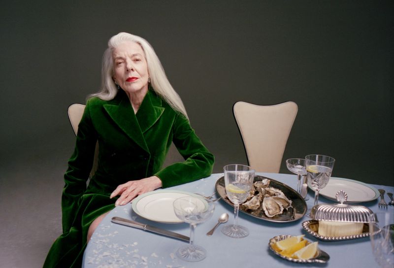 Elderly woman with long white hair wearing a green velvet coat seated at a dining table set with oysters and water glasses